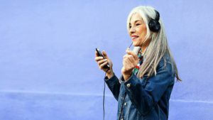 A person with their headphones holding a mobile device and smiling with a purple background