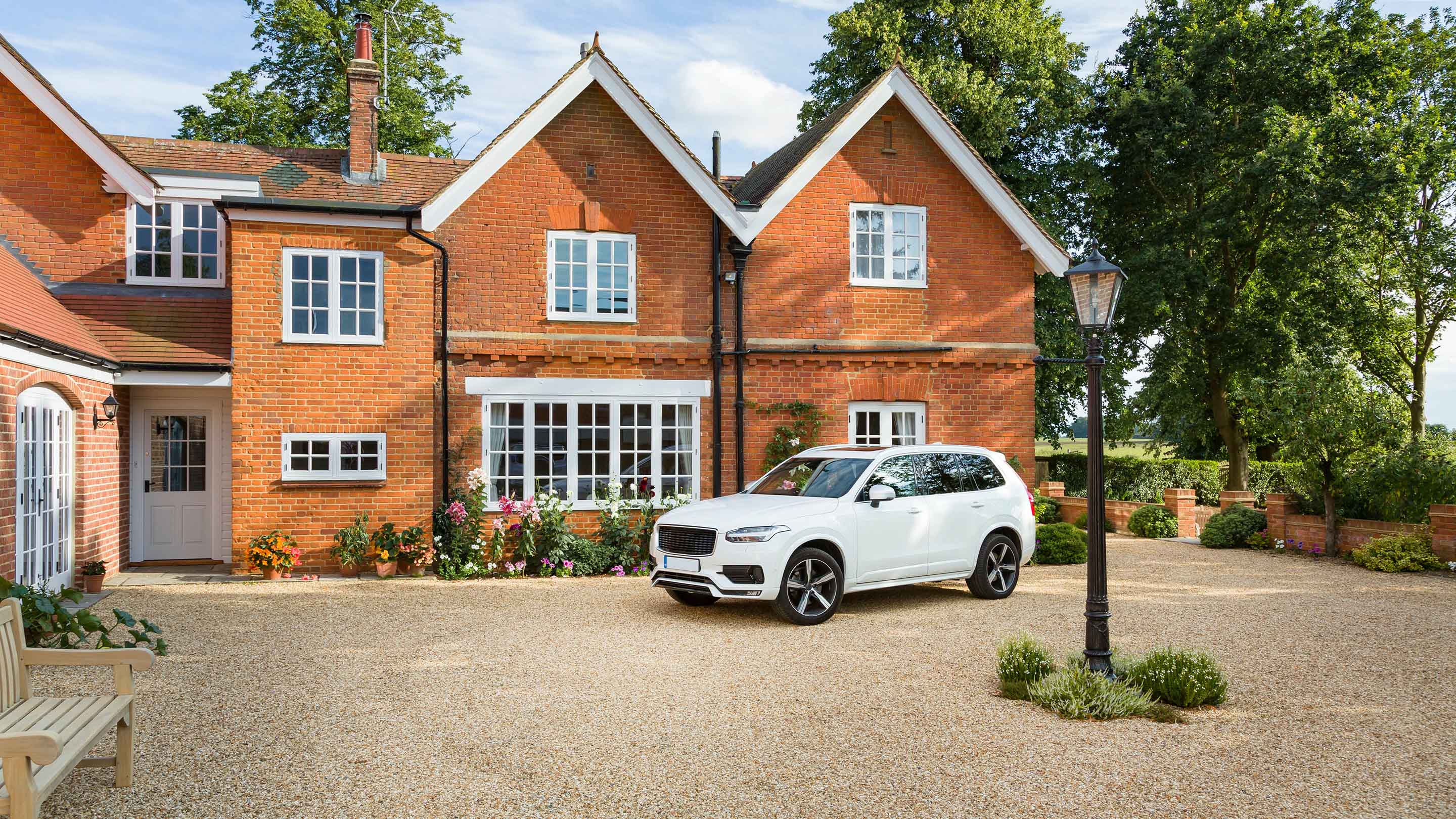A large white car parked outside a house in the sunshine. There are green trees in the background.