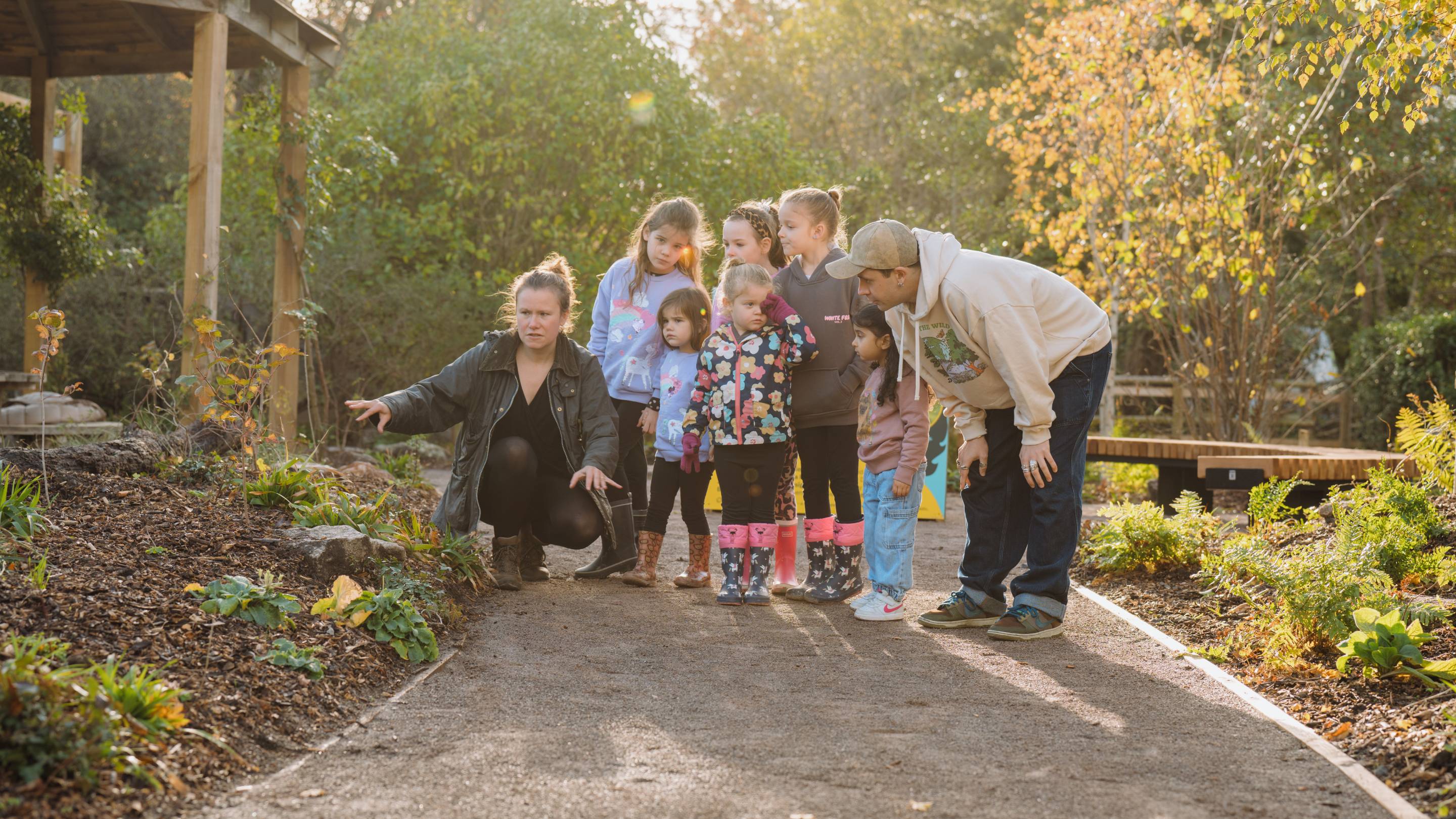 Zoe Claymore showing a group of children and Cel Spellman around the garden path at Bristol Zoo’s temperate rainforest garden. The scene features autumn foliage, green plants, and a wooden bench in the background, with sunlight filtering through the trees.