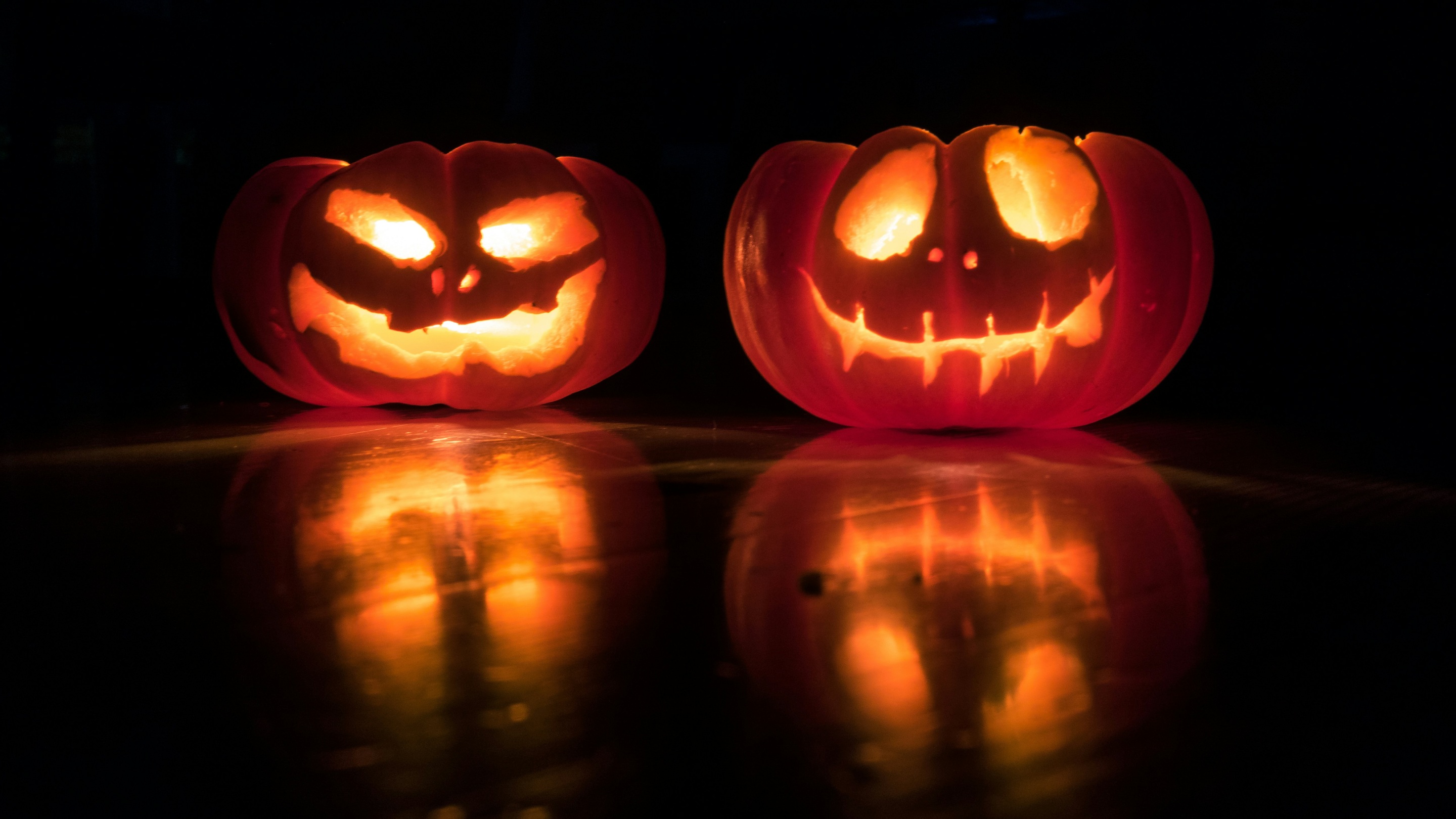Two glowing jack-o’-lanterns with carved faces, lit from inside and reflected on a dark surface