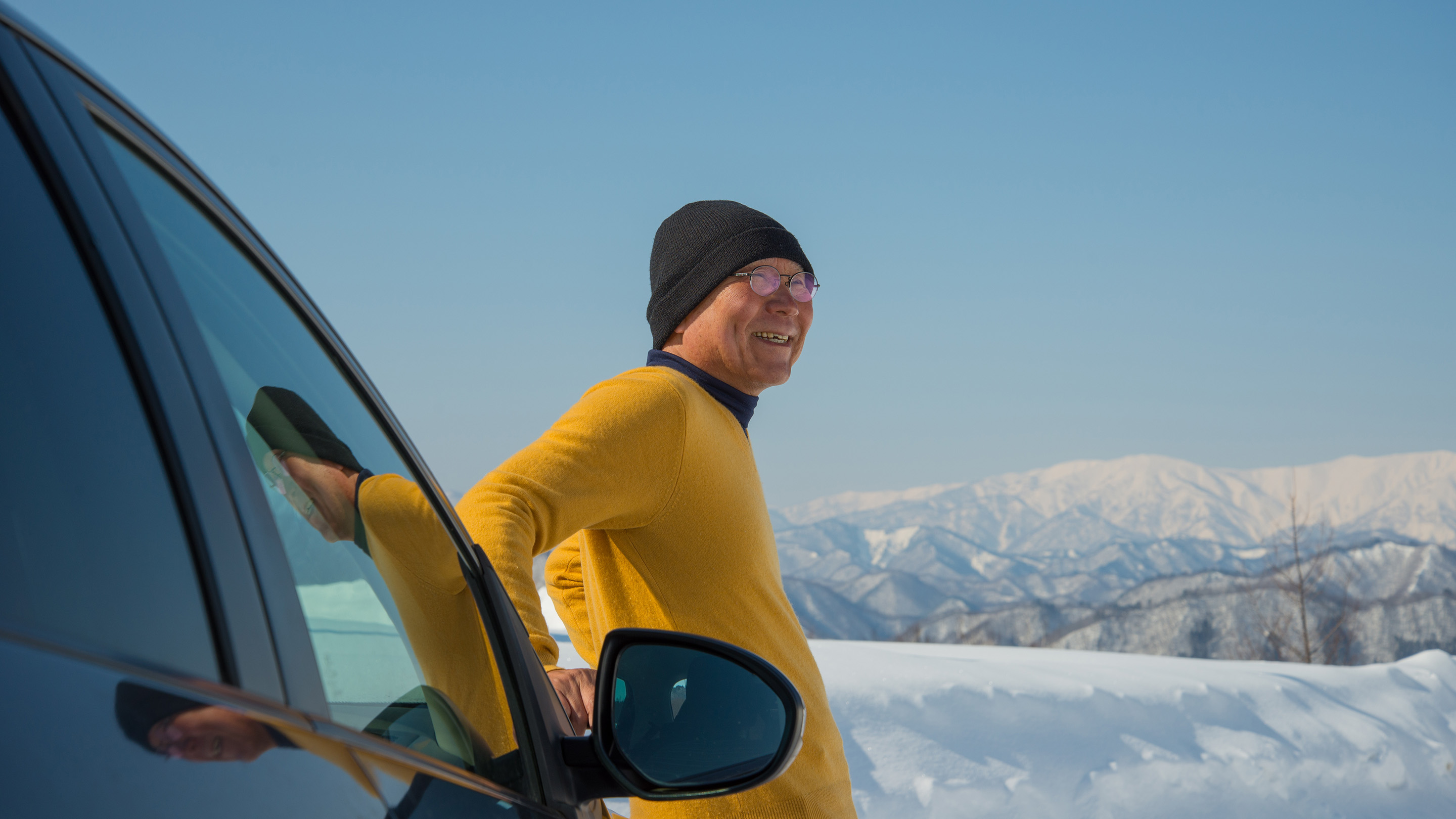 Man standing next to his car, surrounded by snowy mountains.