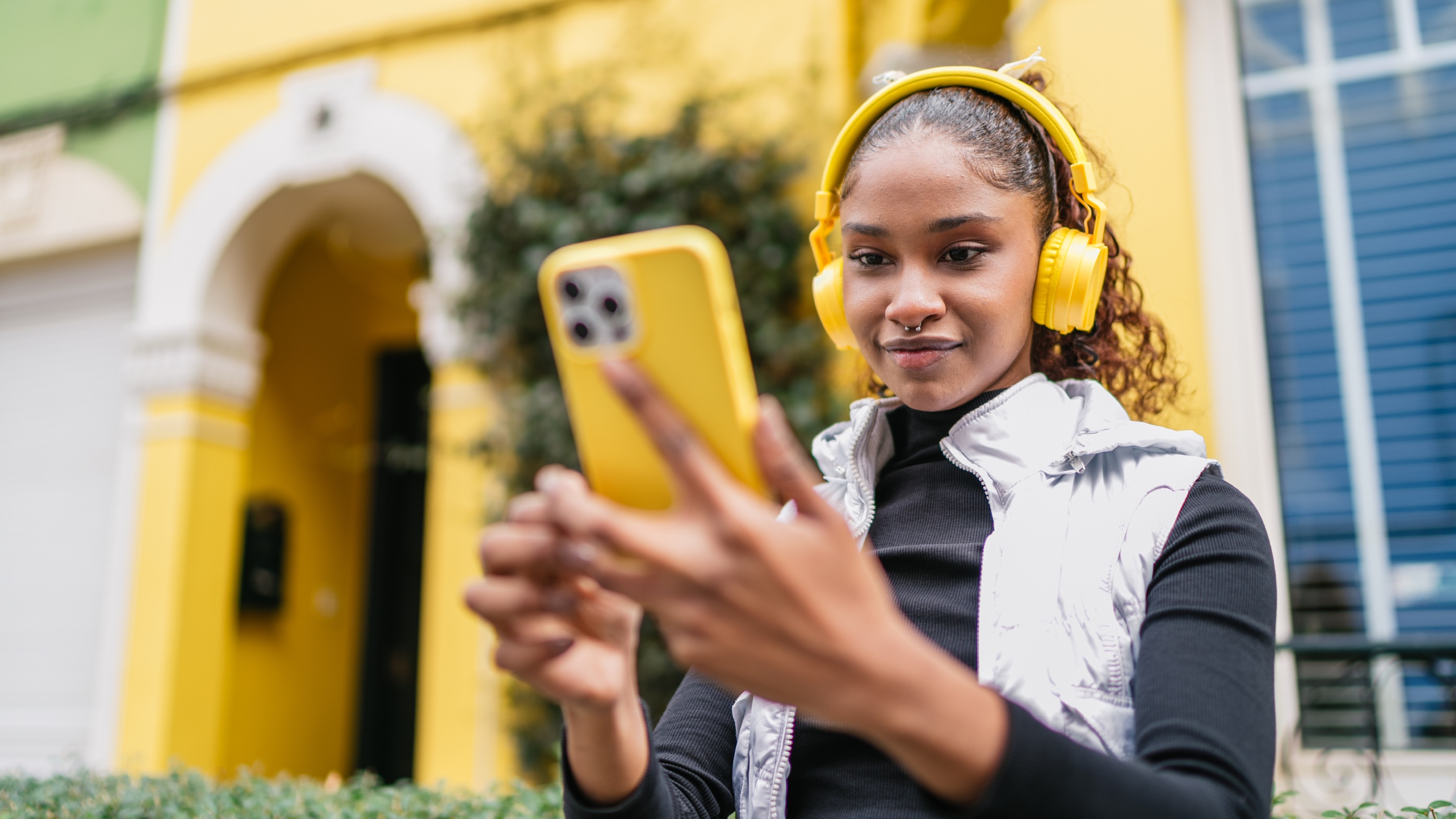 A person wearing yellow headphones and a silver vest is using a yellow smartphone. The background features a vibrant yellow building with an arched doorway and green foliage. The person appears to be focused on the smartphone screen while standing outdoors.