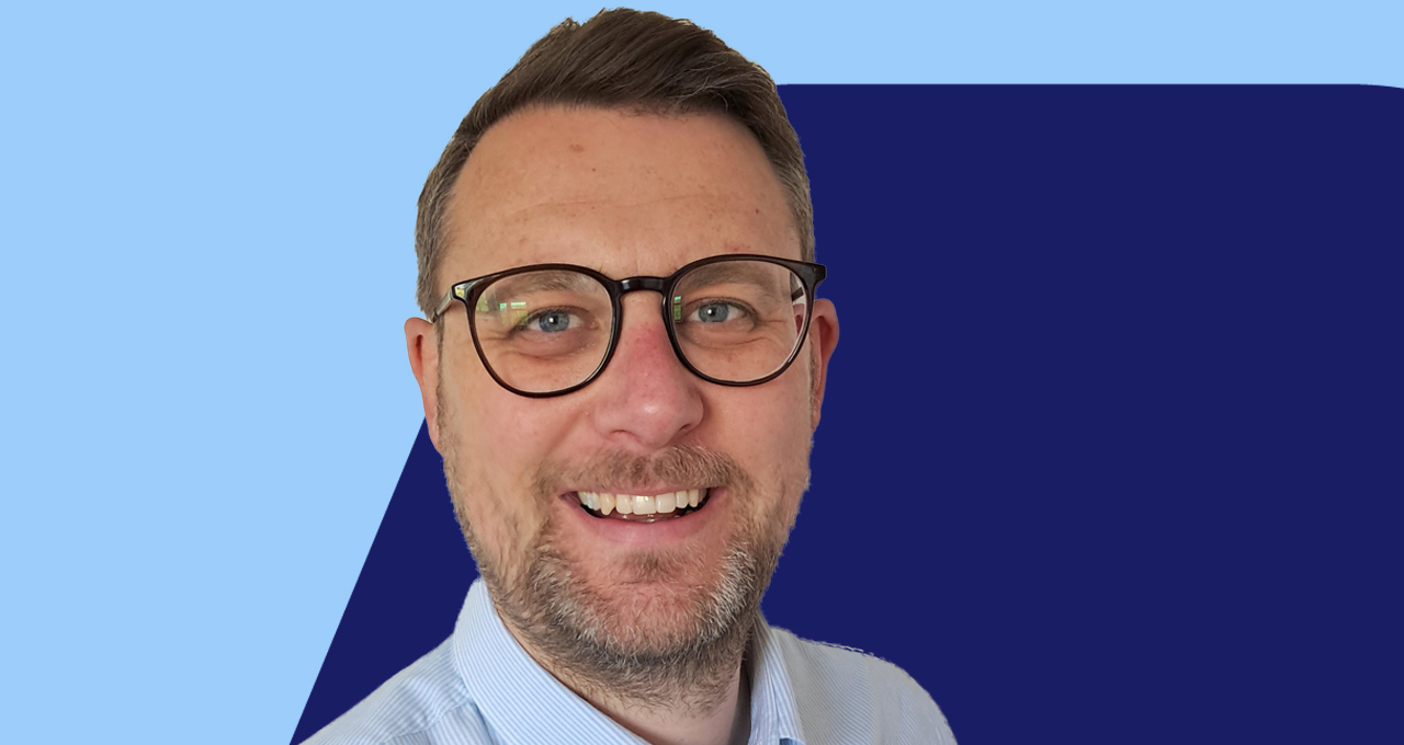 Headshot of Dave Wheately, dark haired male wearing glasses on blue background