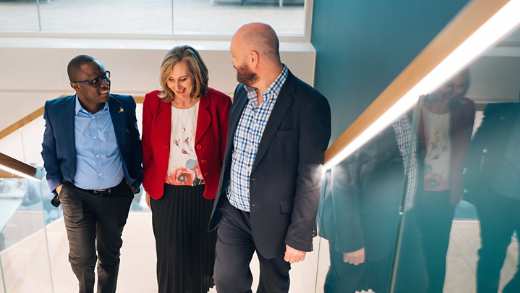 Three professionals of various genders and ethnicities are walking up some stairs, they are smiling whilst talking to wach other