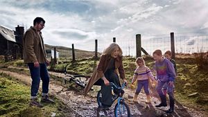 Grandparent playfully riding a child's bicycle down a stoney entrance to a remote countryside house, sourrounded by their adult child and grandchildren.