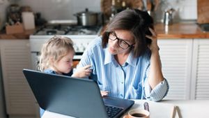 A parent with a strained expression on their face, trying to do something on their laptop while their child is trying to get their attention, in an indoor setting.