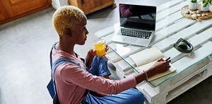 Person sat at a table, drinking juice, whilst writing in their notepad, in a home setting