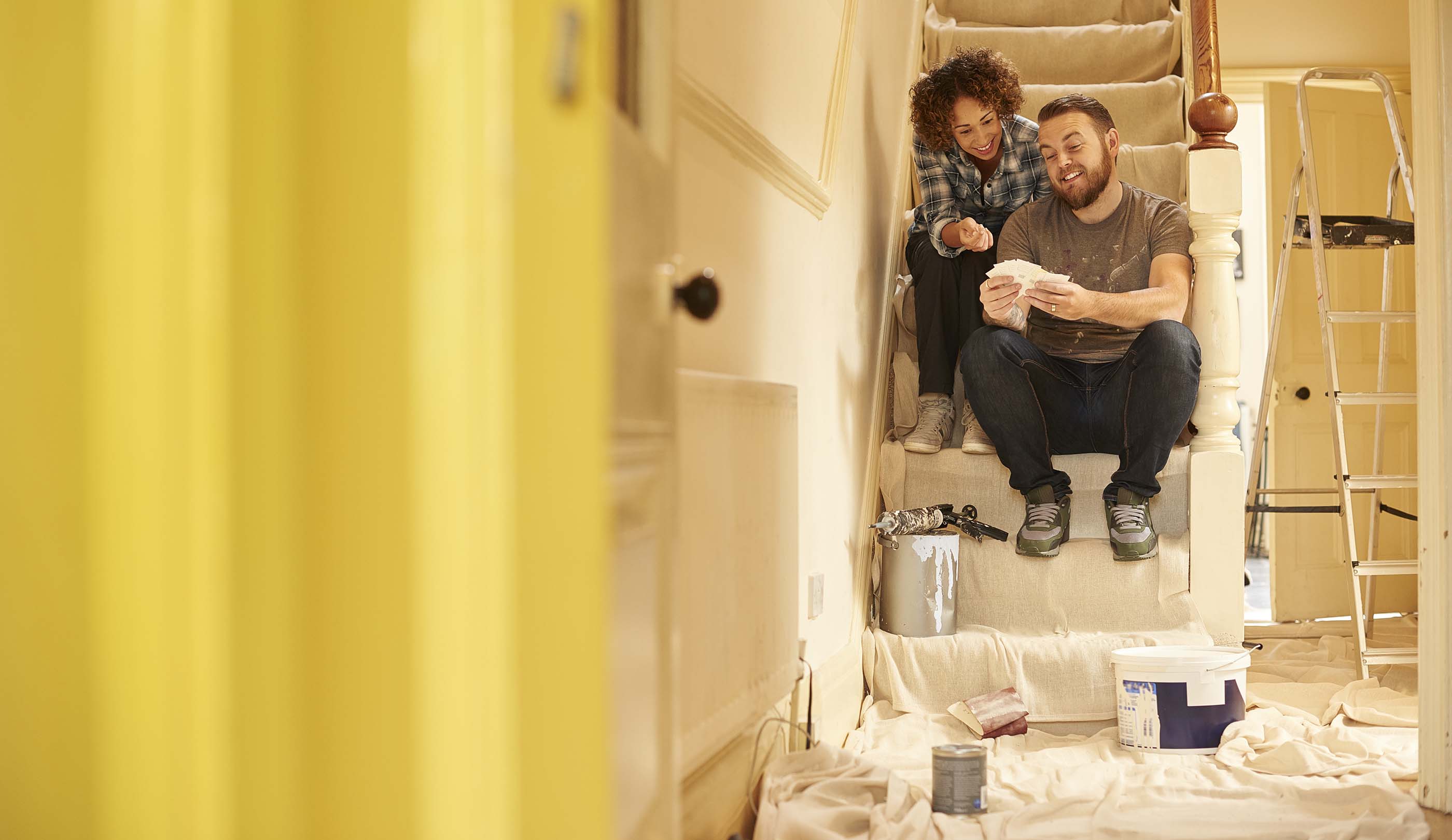Two people sat on a staircase looking at paint swatches. There is a sheet on the ground and cans of paint. They are in the middle of decorating.