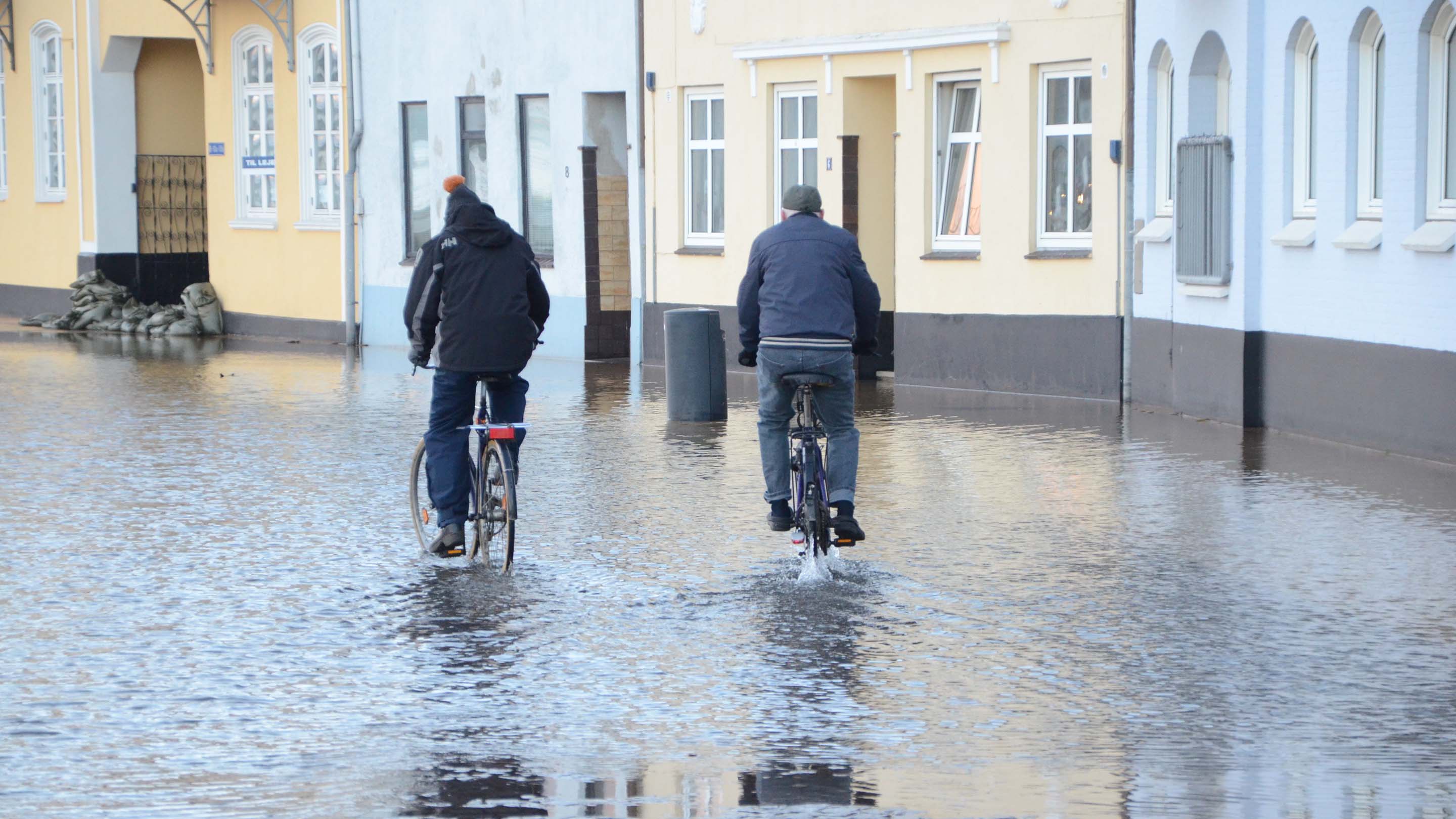 Two men riding bikes through flood water in a town.