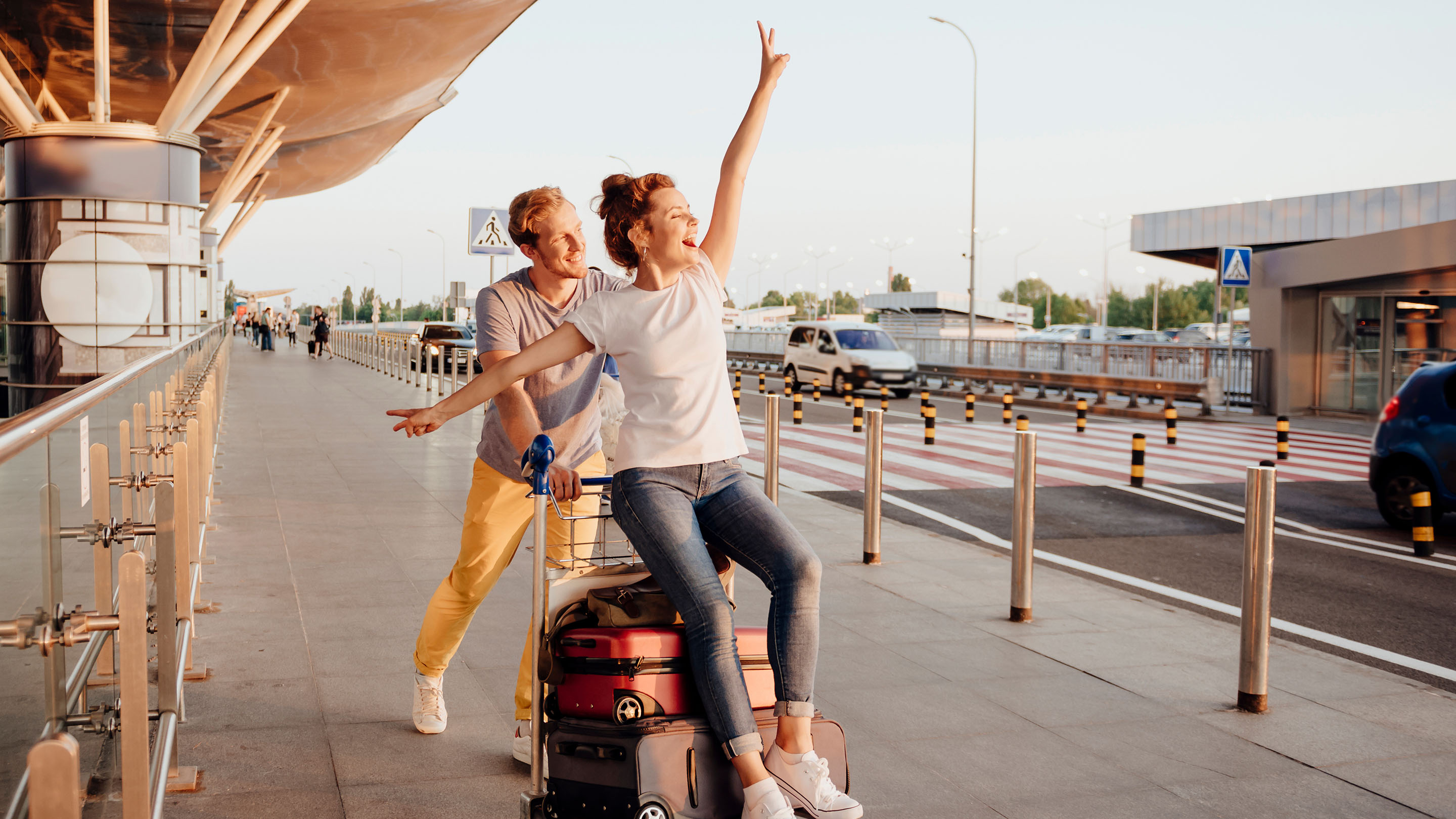Man and woman outside an airport. The woman is riding on top of a luggage cart while the man pushes it.