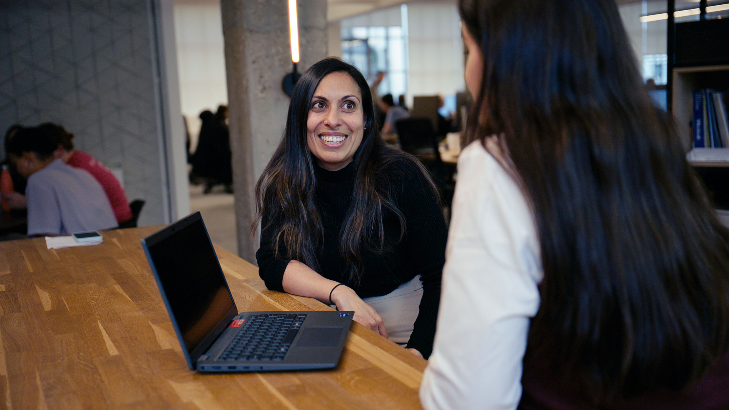 Aneesa sitting at a table, talking to someone with a laptop. Aneesa is smiling. In the background, people are working and talking.