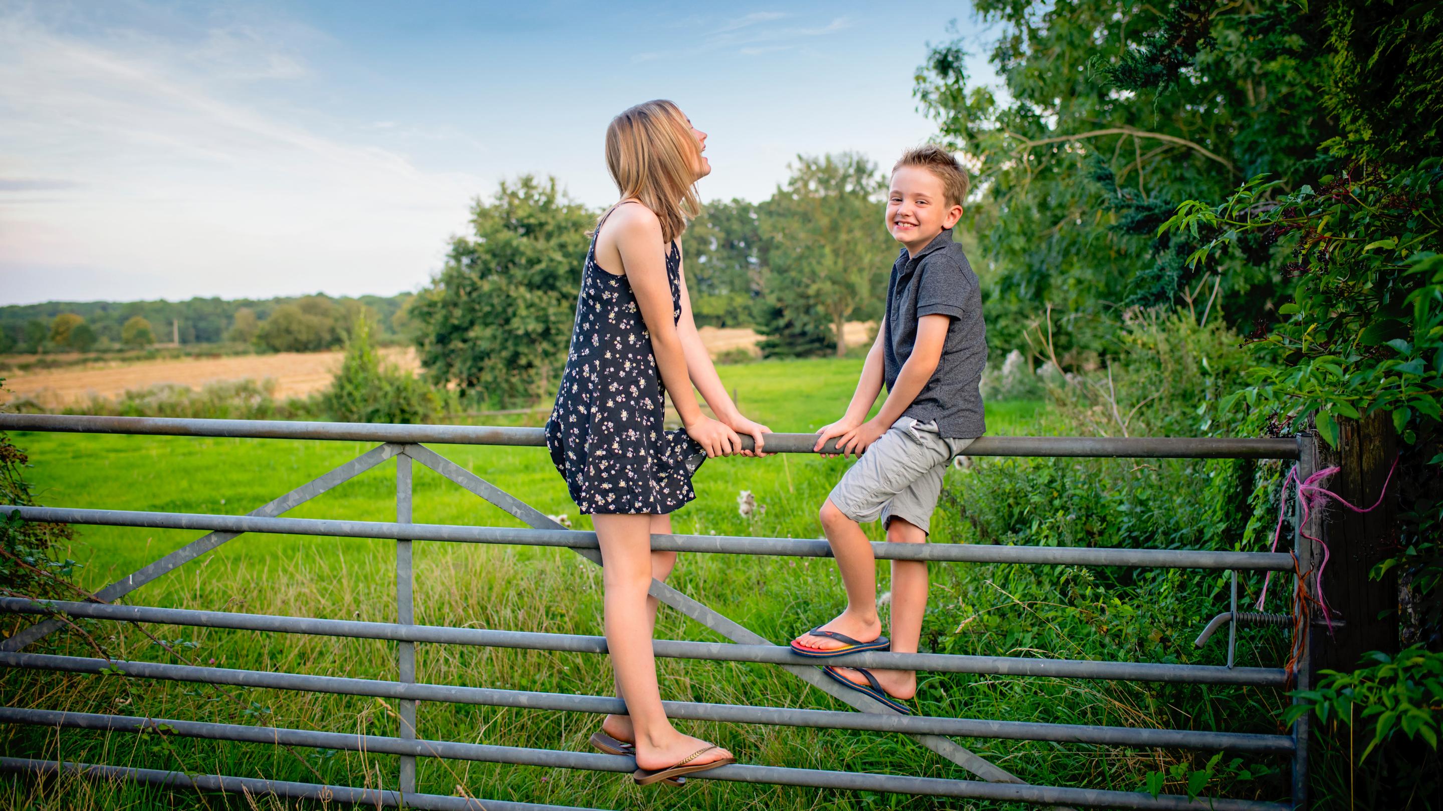 Two children sitting on a metal farm gate in a rural countryside setting, surrounded by green fields, trees and hedgerows on a clear day.