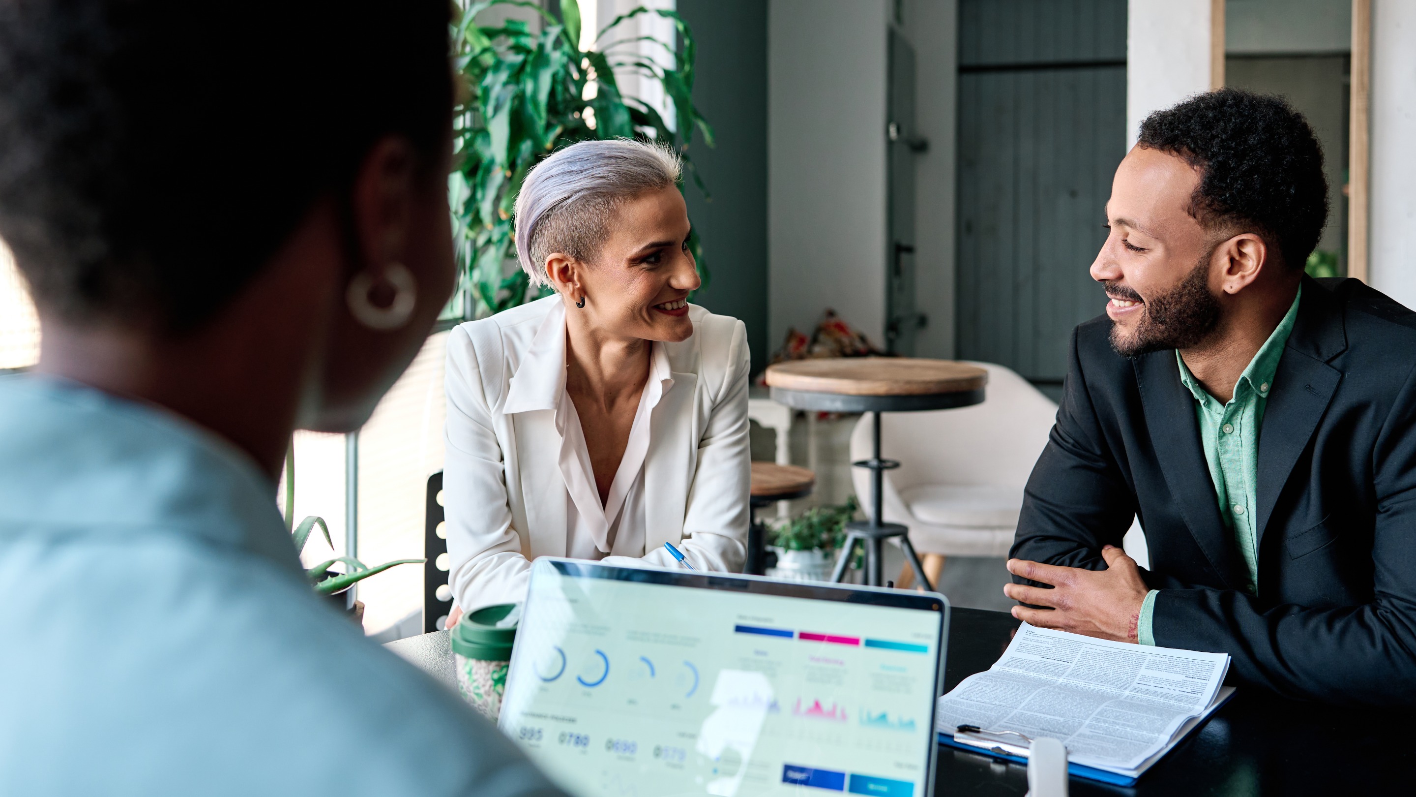 People meeting with clients around a table in a modern office, reviewing documents and business performance data displayed on a laptop.