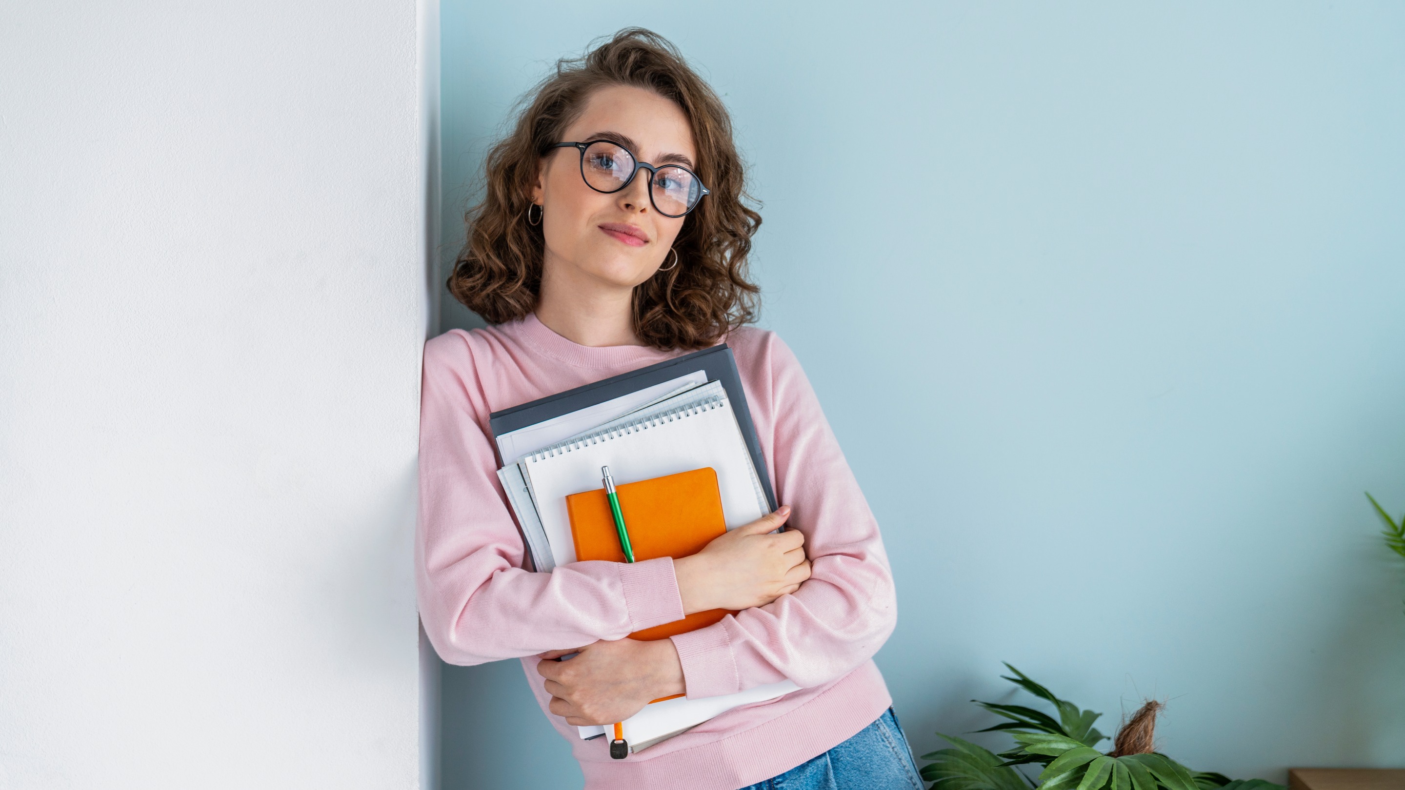 Confident woman holding books, notebooks and pens whilst leaning against a wall