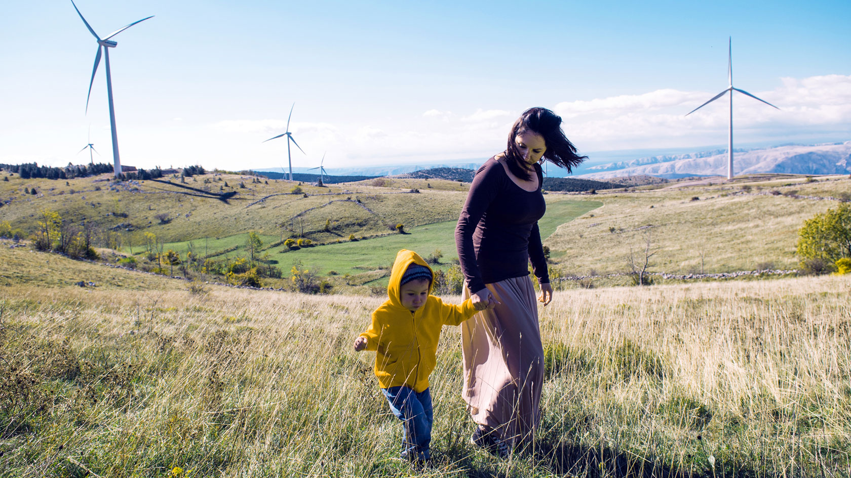 Image of person holding hands with a child, walking across a field with windmills in the background