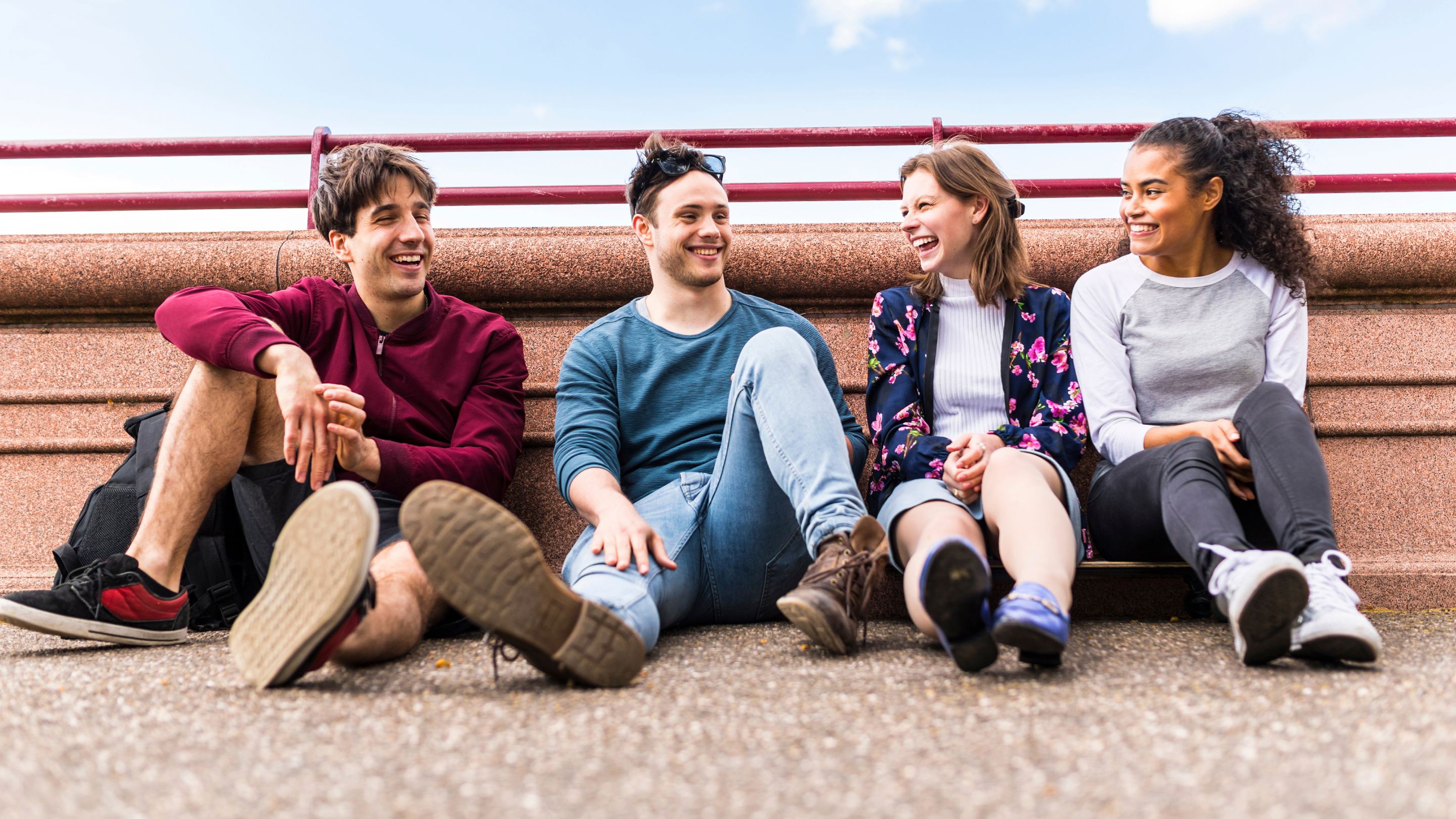 Four people sitting on the ground outdoors, leaning against a stone wall with a red railing, casually dressed and engaged in conversation.