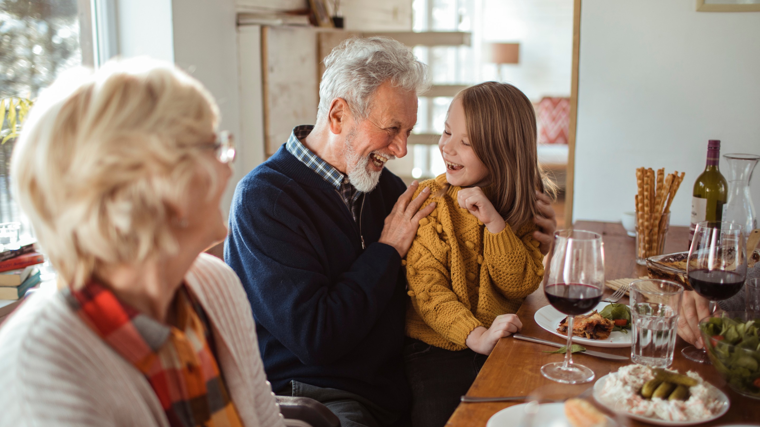 An older adult with grey hair and beard sits at a dining table, smiling and leaning toward a young child in a mustard jumper, while another older adult is blurred in the foreground. The table is set with plates of food, wine glasses, and breadsticks, creating a warm, home setting.