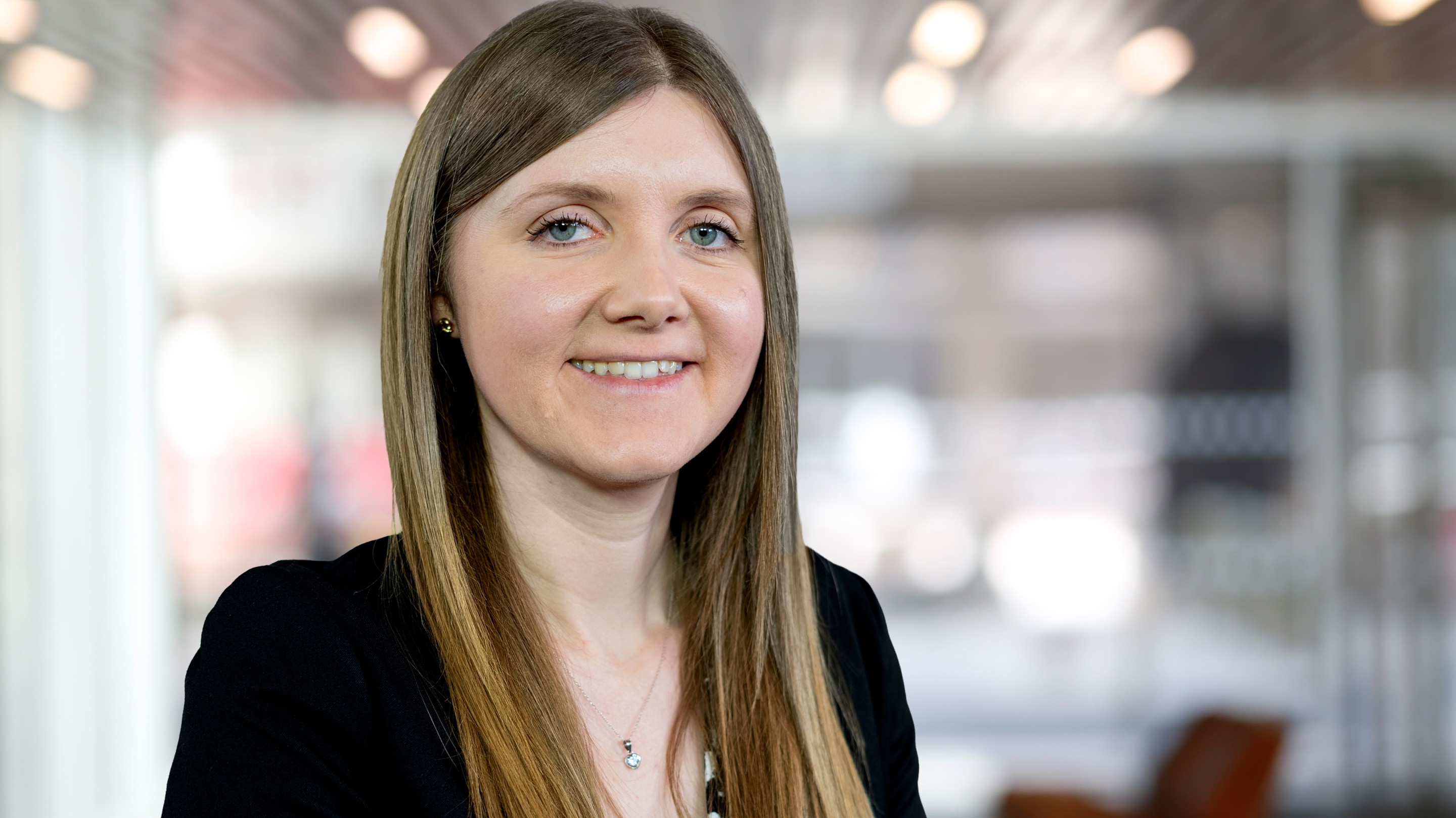 Professional head-and-shoulders portrait of a person with long straight hair, wearing a dark blazer and necklace, looking towards the camera against a softly blurred office background.