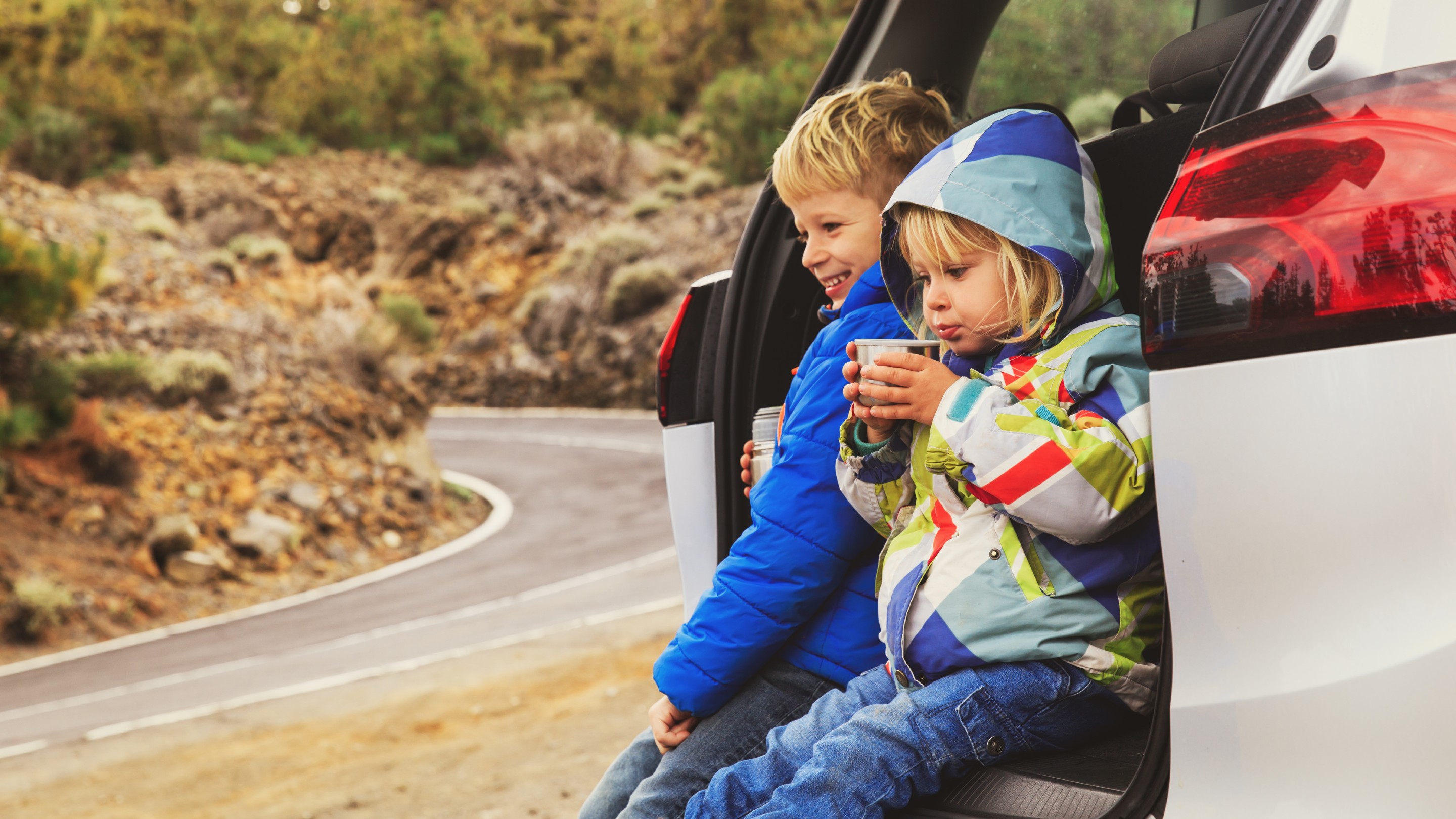 Little boy and girl sit in an open car boot on the side of the road