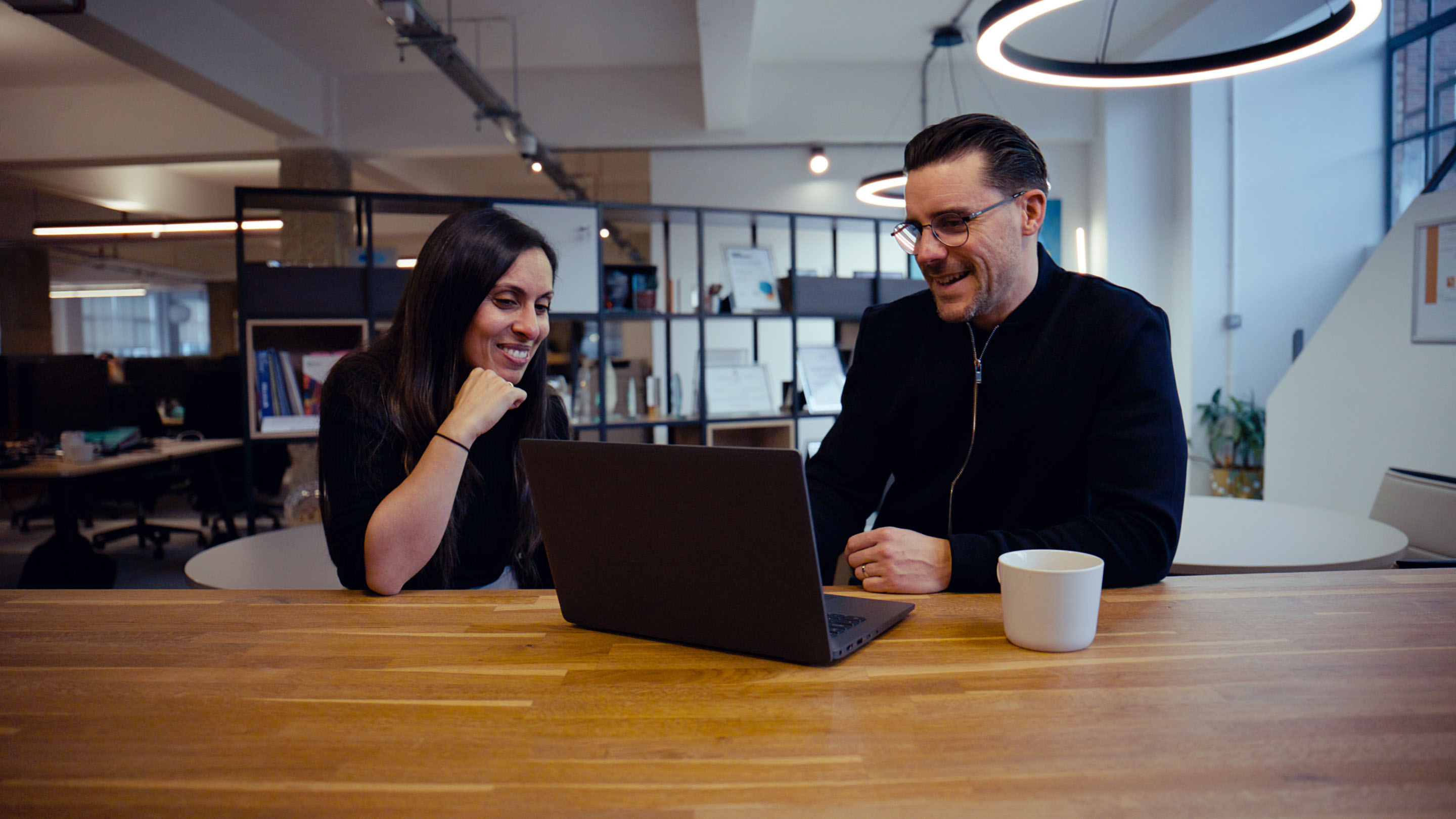 Luke and Aneesa talk in Mencap's office. They're sitting in the kitchen area and smiling while looking at a laptop.