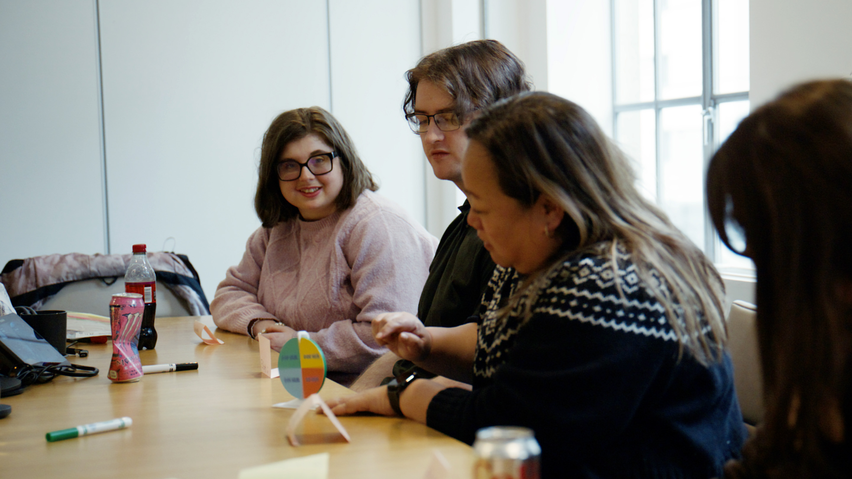 Three people in a co-creation session. They are sat around a table. The person in the foreground is using a 'spinner' with different sums of money on it. The person in the background is smiling.
