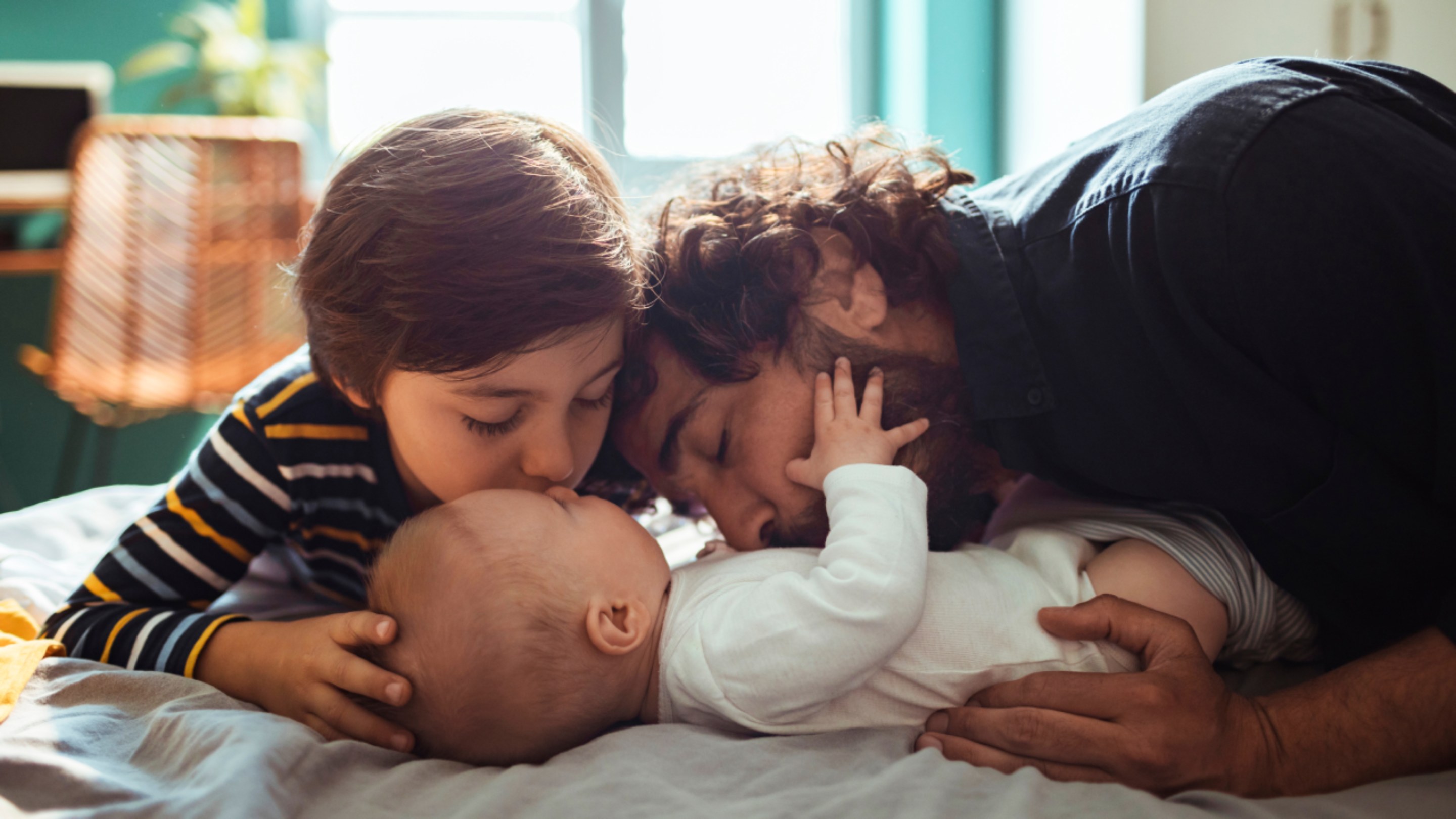 A parent and older child lie together on a bed, affectionately cuddling and kissing a baby in a warm family home setting