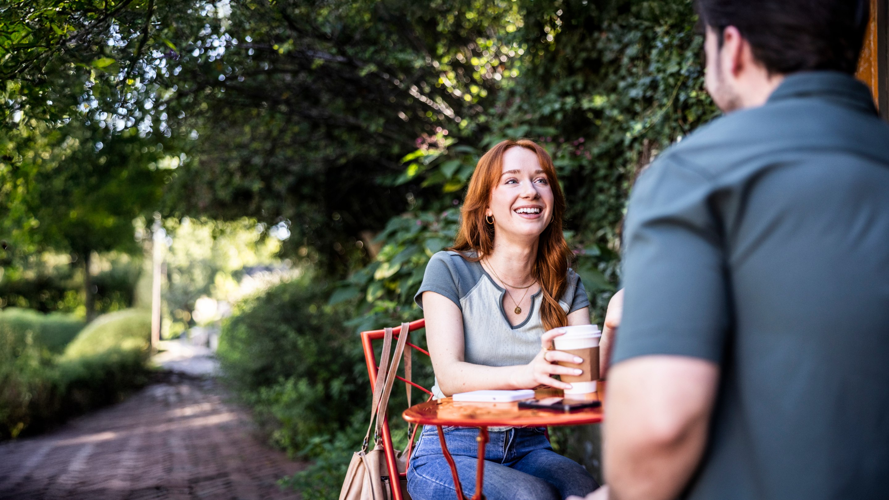 Two people sitting at an outdoor cafe table having a conversation over coffee on a leafy garden path.