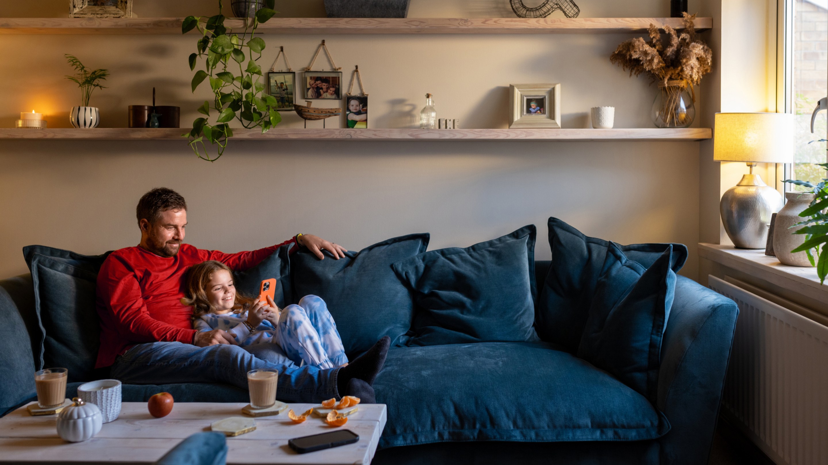An adult and a child sit together on a blue sofa in a living room, looking at a smartphone, with drinks and fruit on a coffee table and family photos displayed on shelves behind them.