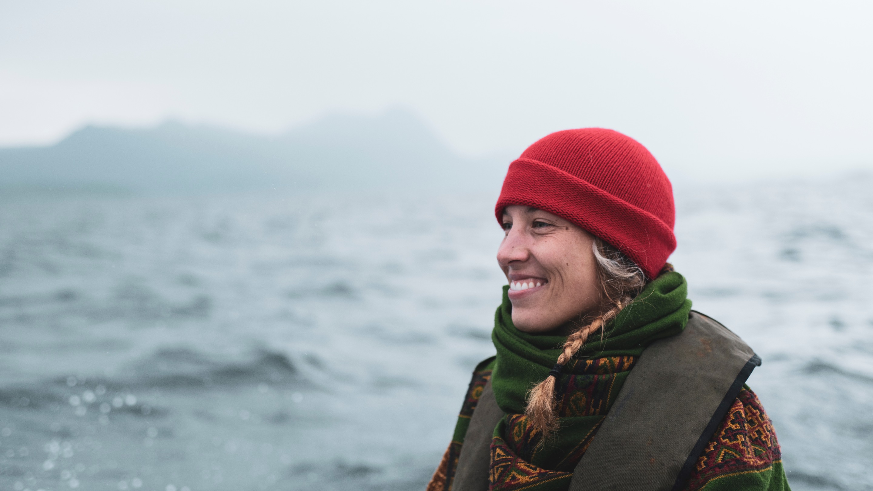 A person wearing a red beanie and green scarf sits on a boat with choppy grey water and misty mountains in the background.