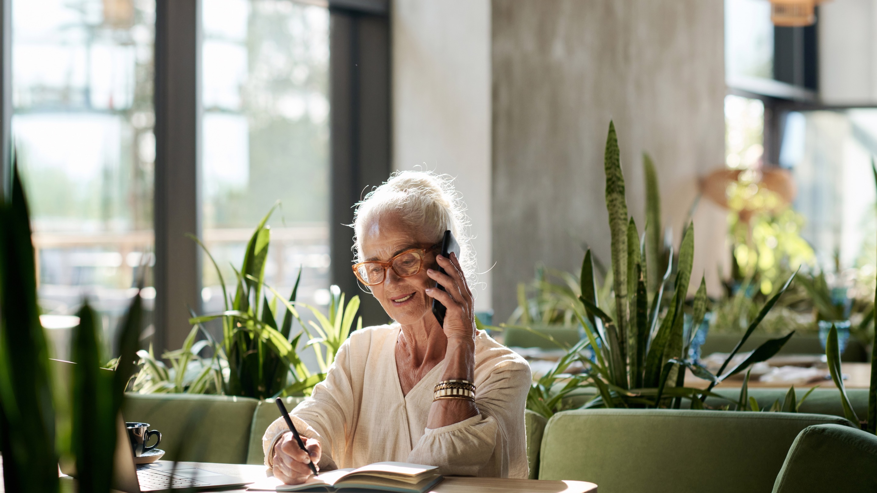 Person sitting at a wooden table in a bright, modern café surrounded by green plants, writing in a notebook while holding a phone, with a laptop nearby.
