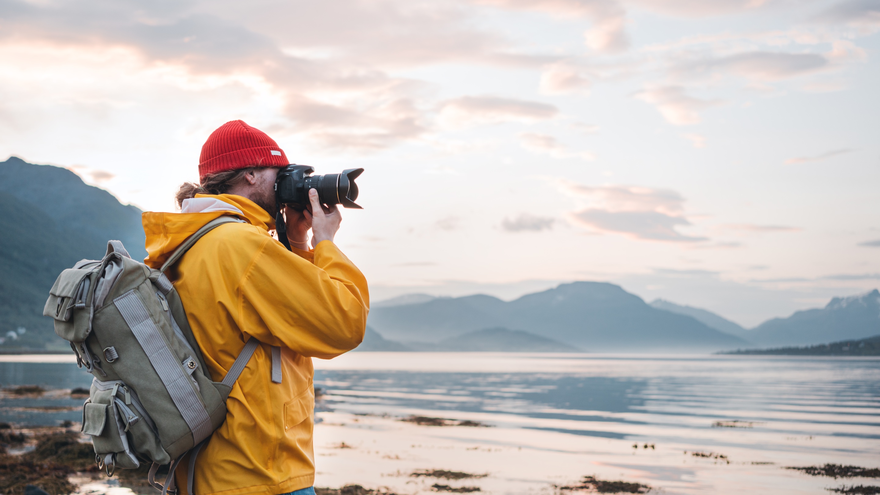 A person wearing a yellow jacket and carrying a backpack uses a camera to photograph a calm lake surrounded by mountains under a cloudy sky.