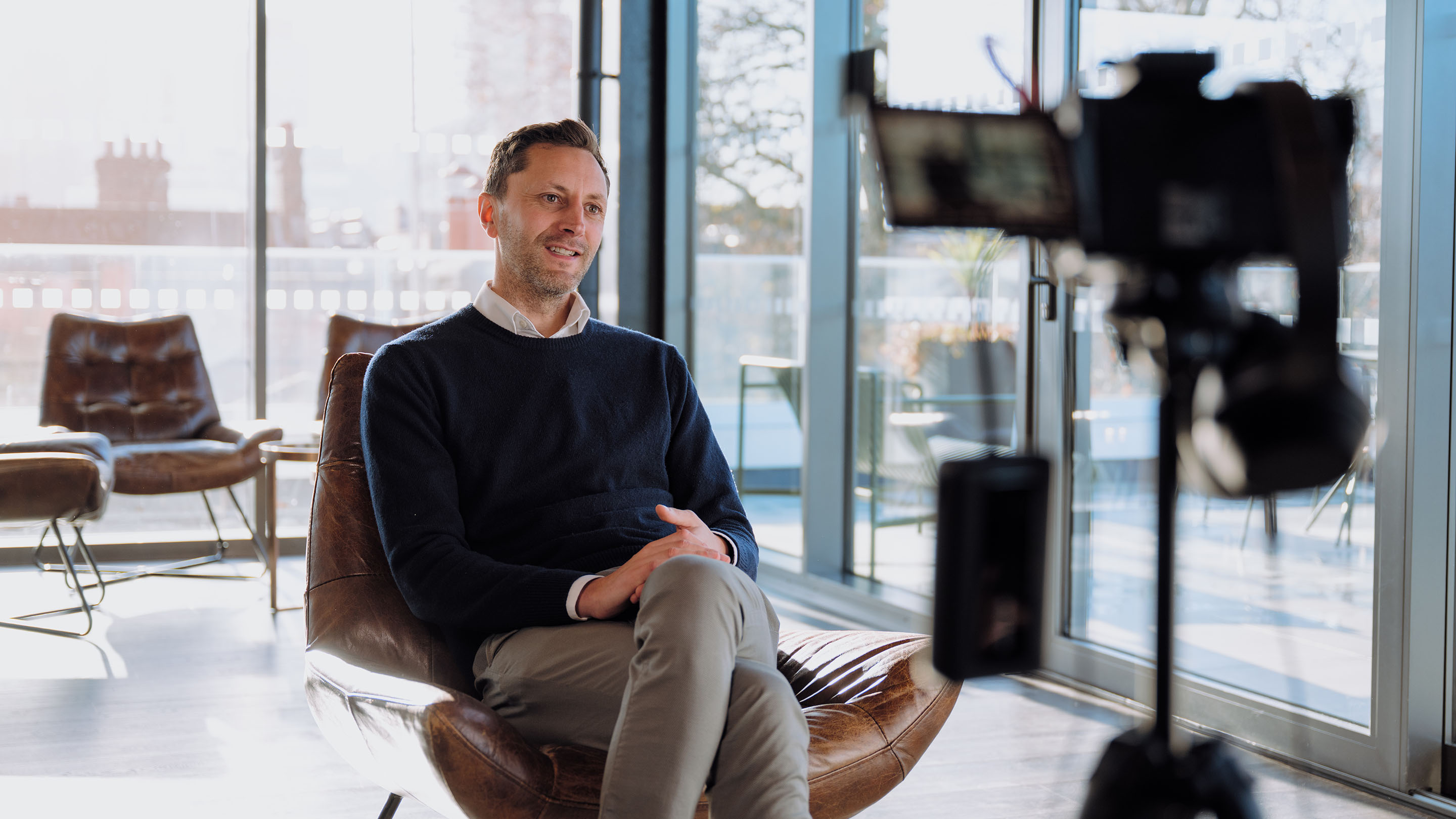 Alex, Aviva Foundation lead, sits down for an interview with us in Aviva's Hoxton office. Alex is sat in a leather chair with bright sunshine coming through the windows behind him. In the blurred foreground is a camera setup pointing at him.
