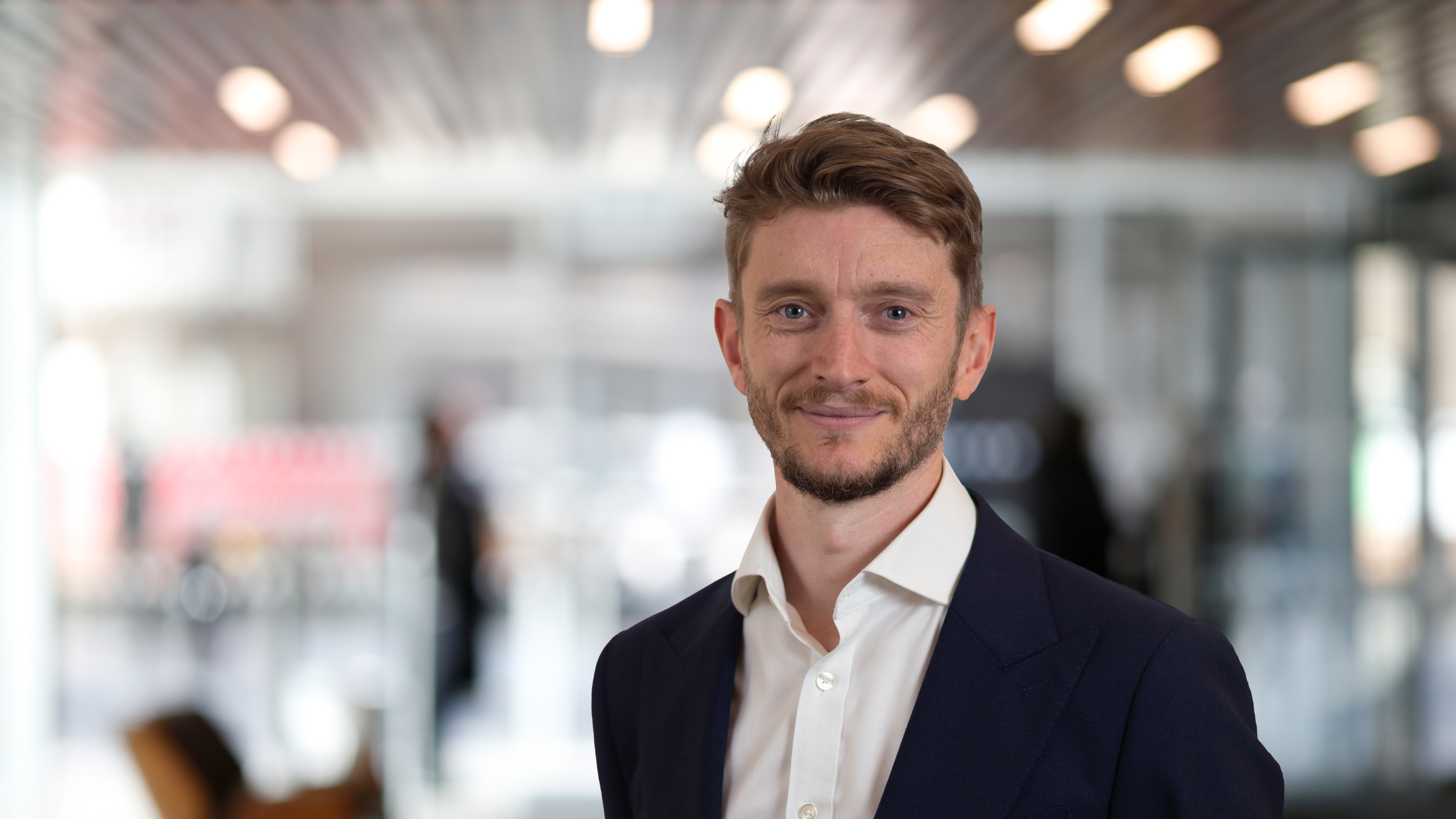 Professional headshot of a person in business attire standing in a modern office with a blurred background