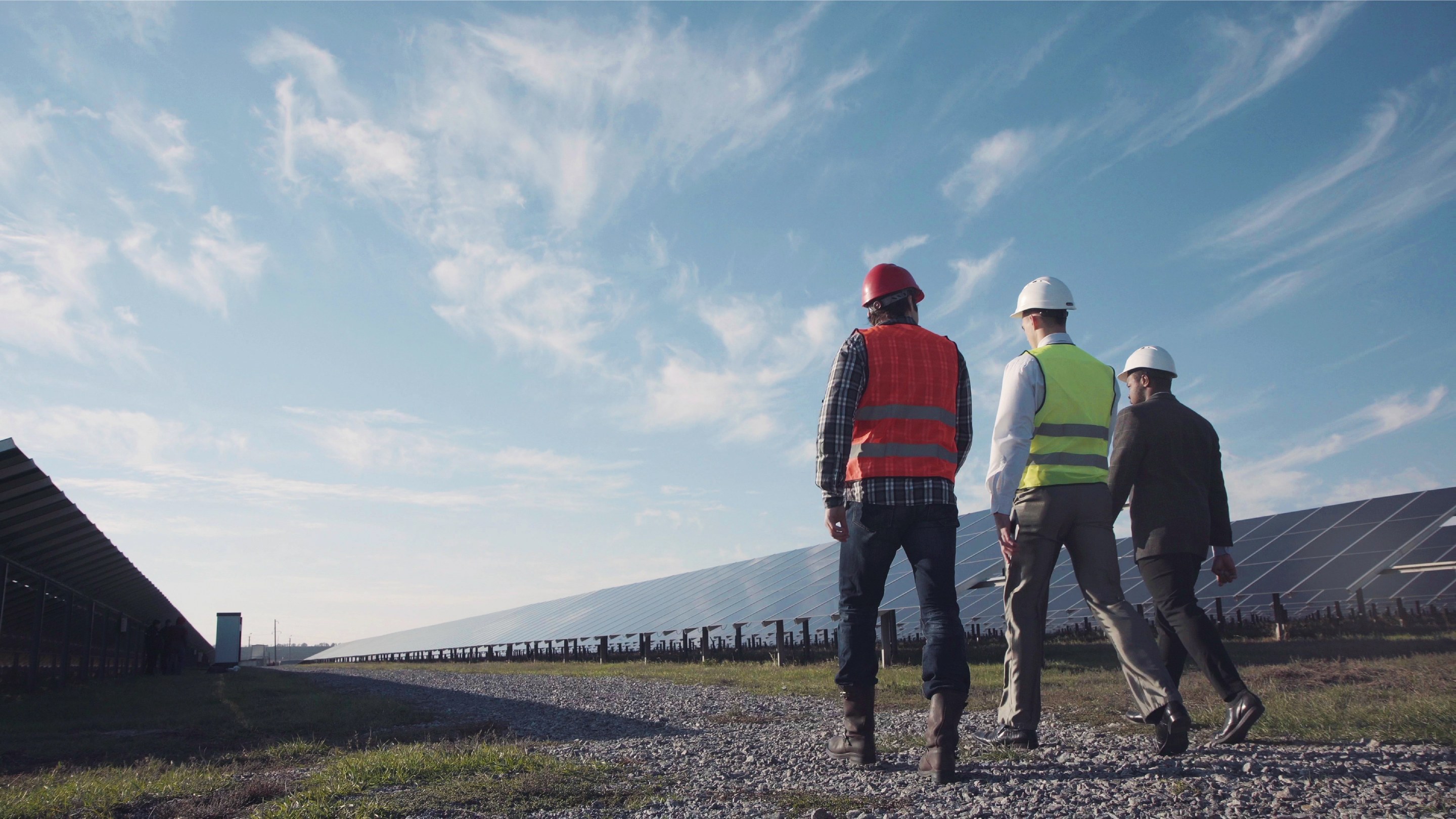 People wearing safety helmets and high‑visibility vests walk through a solar energy site with rows of solar panels under a blue sky.
