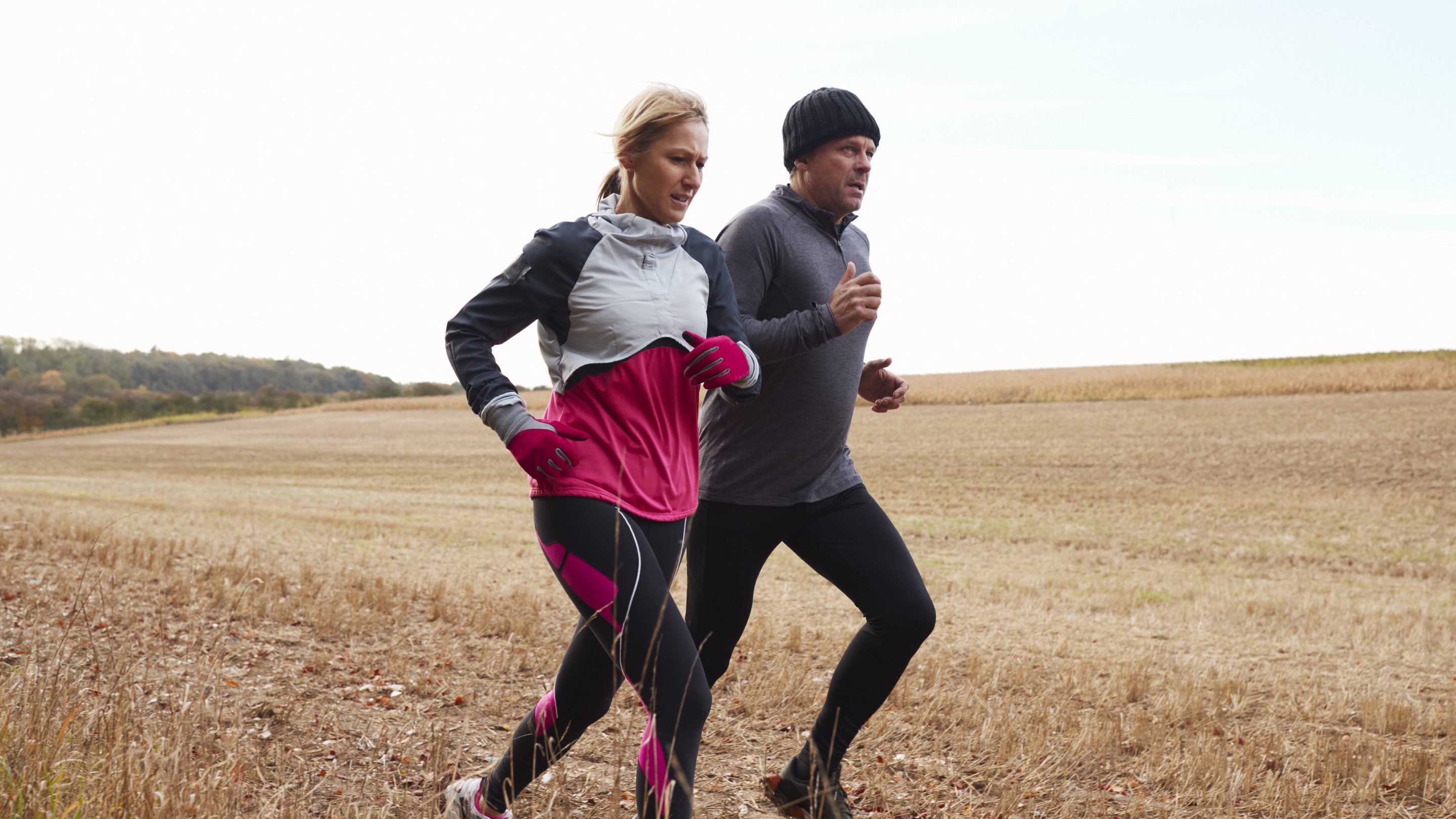Two people running together along a dry, open field on a clear day, wearing athletic clothing.