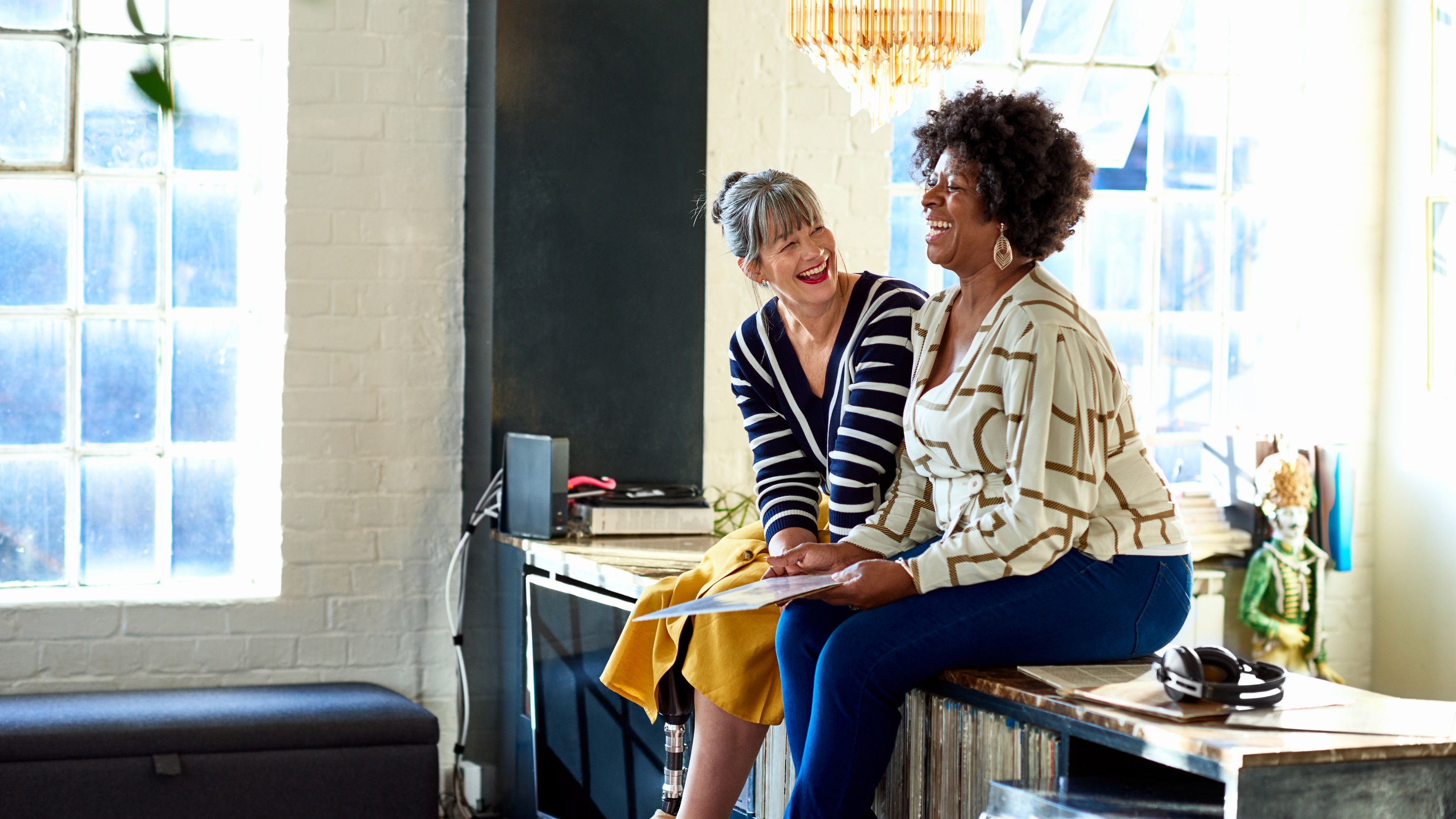 Two people sitting on a cabinet in a bright office space, talking together with natural light from large windows in the background.