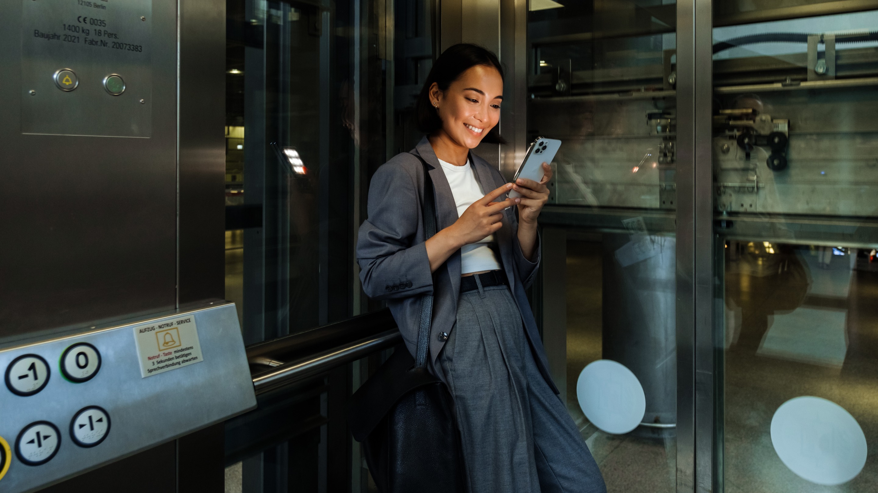 Person standing inside a glass lift, leaning against the wall and checking a smartphone, with metal panels and control buttons visible around them.