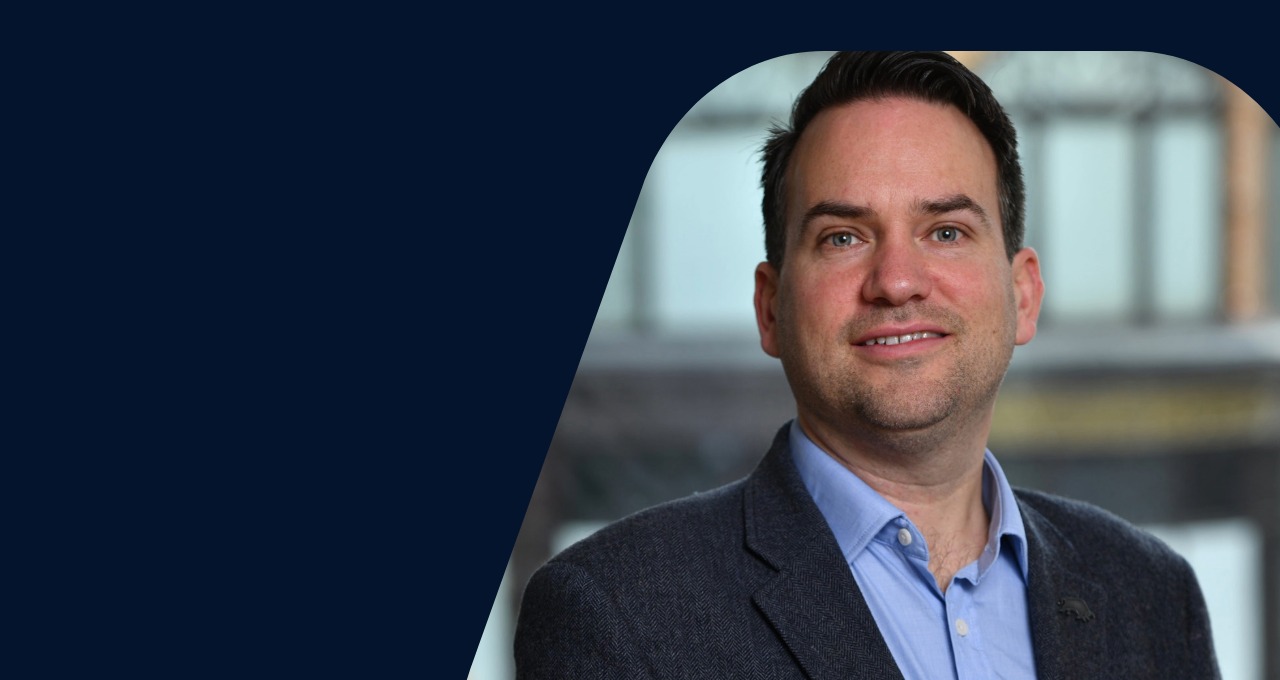 Simon Martin looking to camera and smiling. He wears a blue shirt and a dark jacket.