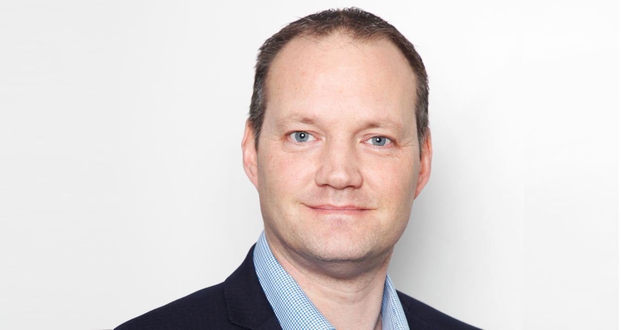 Headshot of Julian Nurse, white male, cropped hair, blue eyes, wearing a suit on pale background