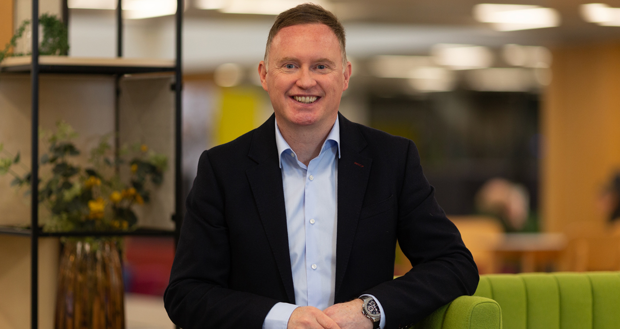 Upper body photo, male in navy suit, pale blue shirt open collar against office backdrop