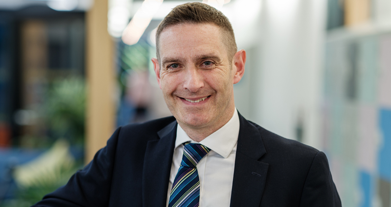 Headshot of Neil Carpenter. Dark haired male in black suit with white shirt and tie, smiling at camera